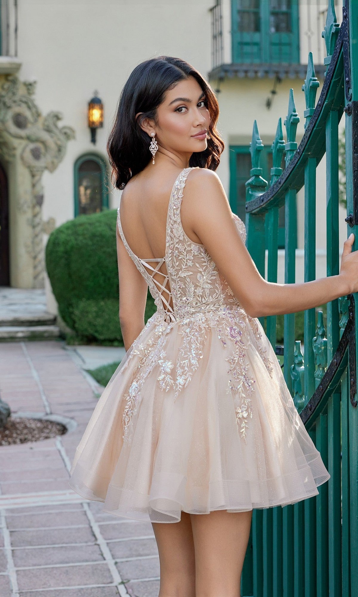 A woman wearing the Juliet JT987N Short Homecoming Dress in champagne, featuring a beautifully embroidered bodice and lace detailing, stands by a green gate outdoors, looking over her shoulder.